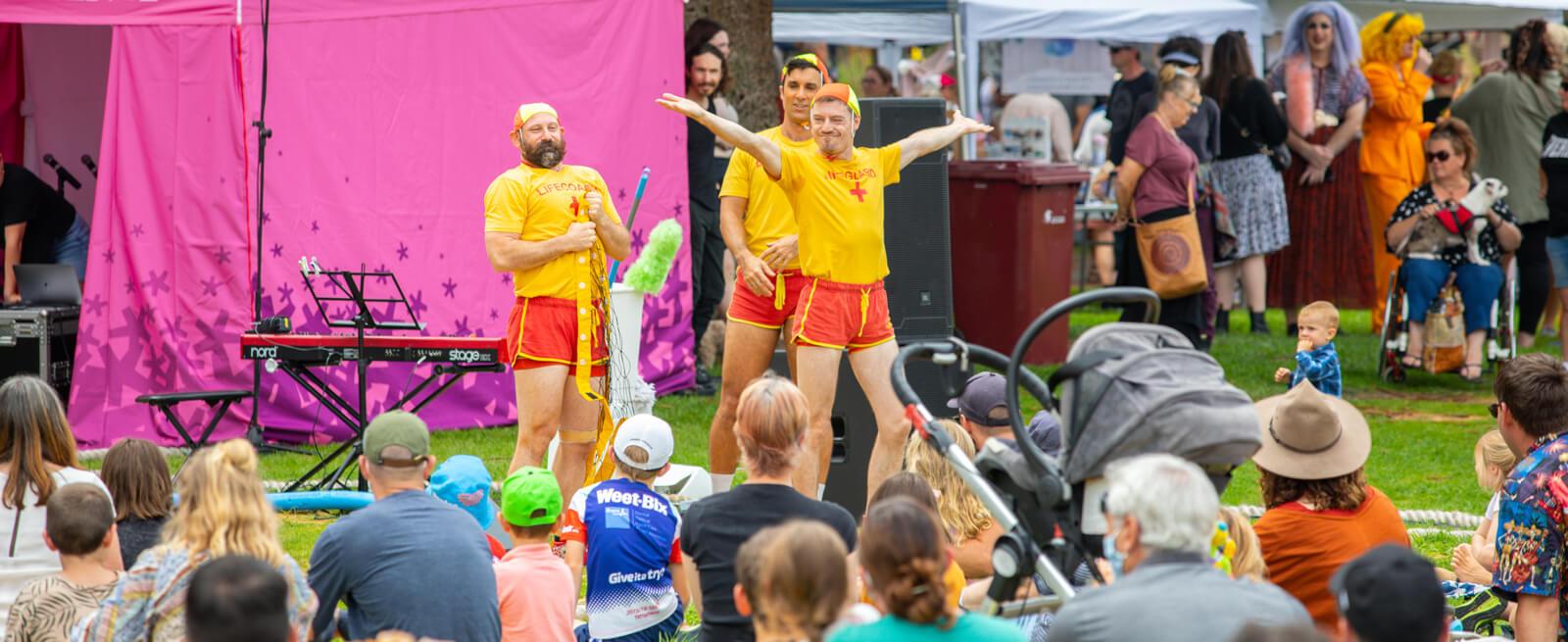 Three happy and celebratory circus artists dressed as lifeguards presenting to a seated audience on grass
