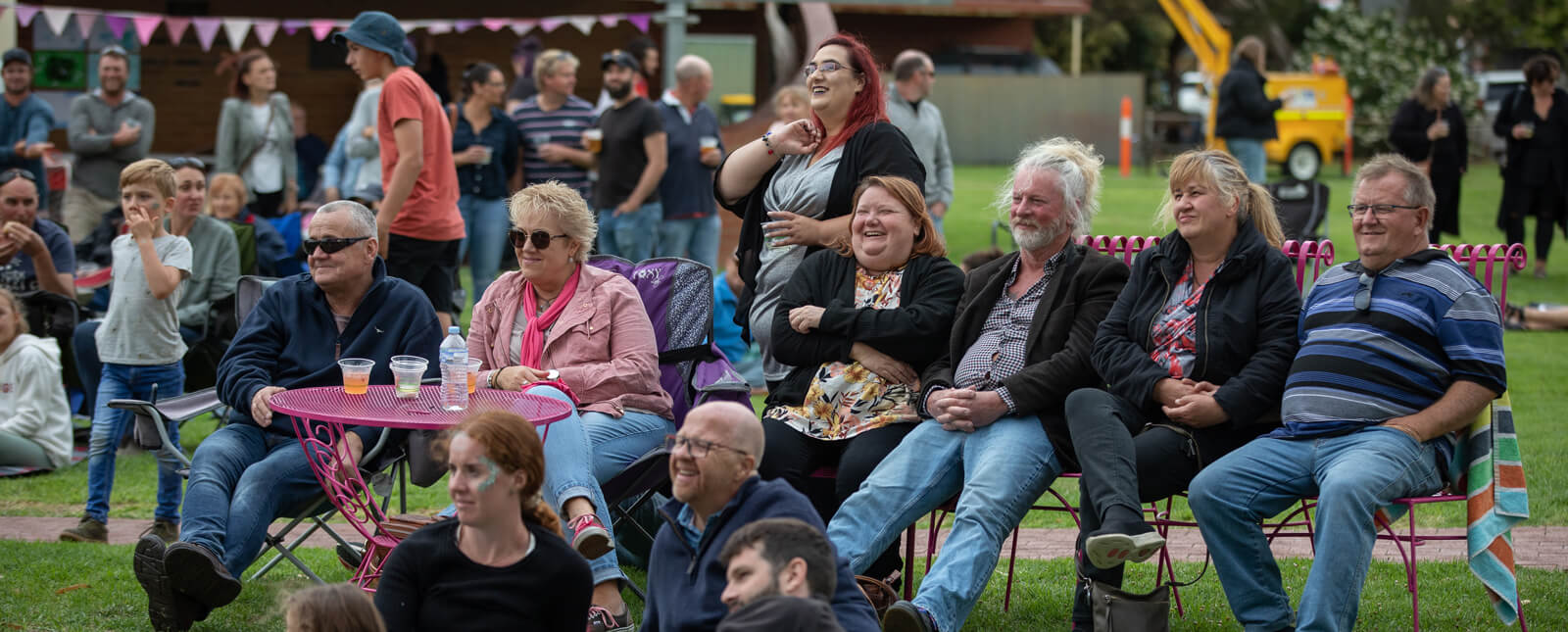 A group of people in laughter, seated on pink metal furniture and picnic blankets in a park at Kingston Fringe