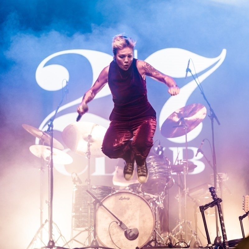 A front woman with a hand-held microphone in a crimson velvet one piece jumps on stage in front of a drum kit and backdrop that reads '27 Club'. 