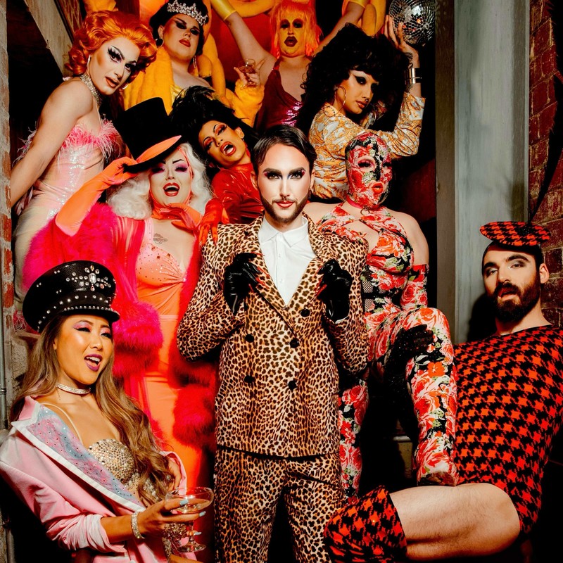 Ten performers in various patterned costumes look toward the camera whilst standing in a stairway 