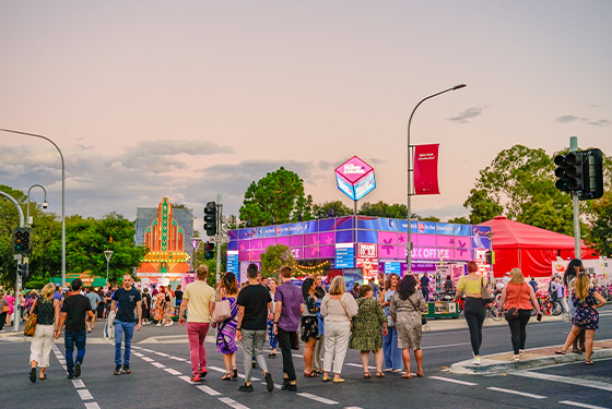 Crowd of people gather and walk across road at twilight in front of purple, pink and blue Adelaide Fringe Corner Box office square structure.  Beside, to the right, a red tent, teetops behind in the distance. 