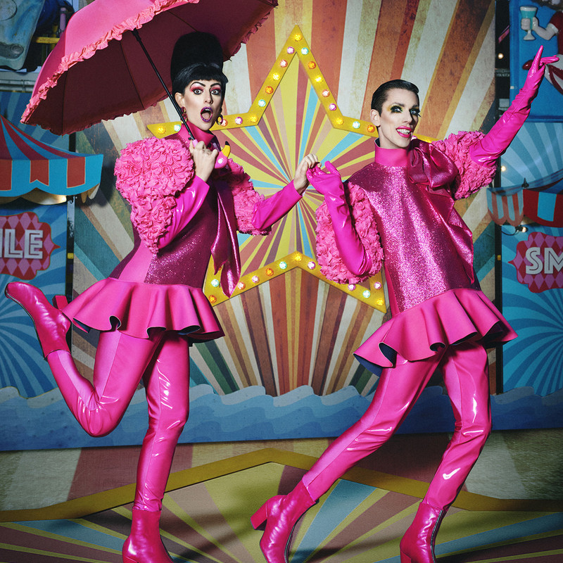 Bourgeois & Maurice wear bright pink outfits made of glitter, frills and leather, posing with a pink umbrella towards the camera 
