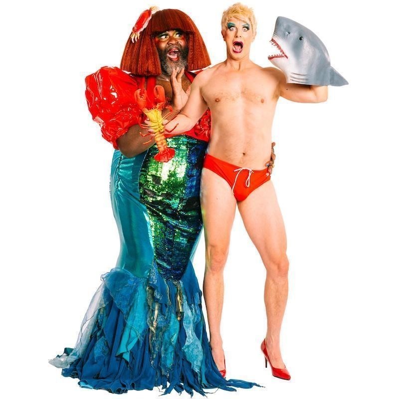 A drag mermaid stands next to a drag artist in a red swimbrief with a shark mask on their hand