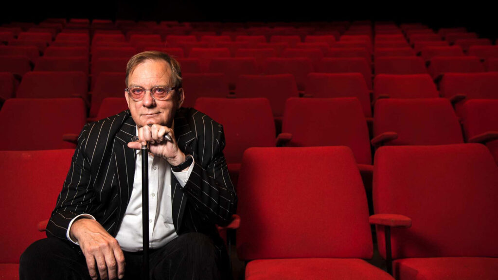 Peter Goers sits in an empty theatre of red chairs, leaning his chin on his walking stick in hand and smirking toward the camera. 