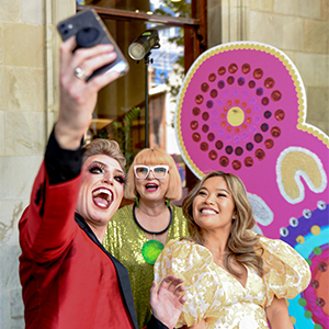 An image of the 2022 Adelaide Fringe Program Launch at The Adelaide Town Hall. The CEO of Adelaide Fringe, Heather Croall and 2022 Fringe ambassadors Reuben Kaye and Diana Nguyen are standing in front of a colourful backdrop taking a selfie. 