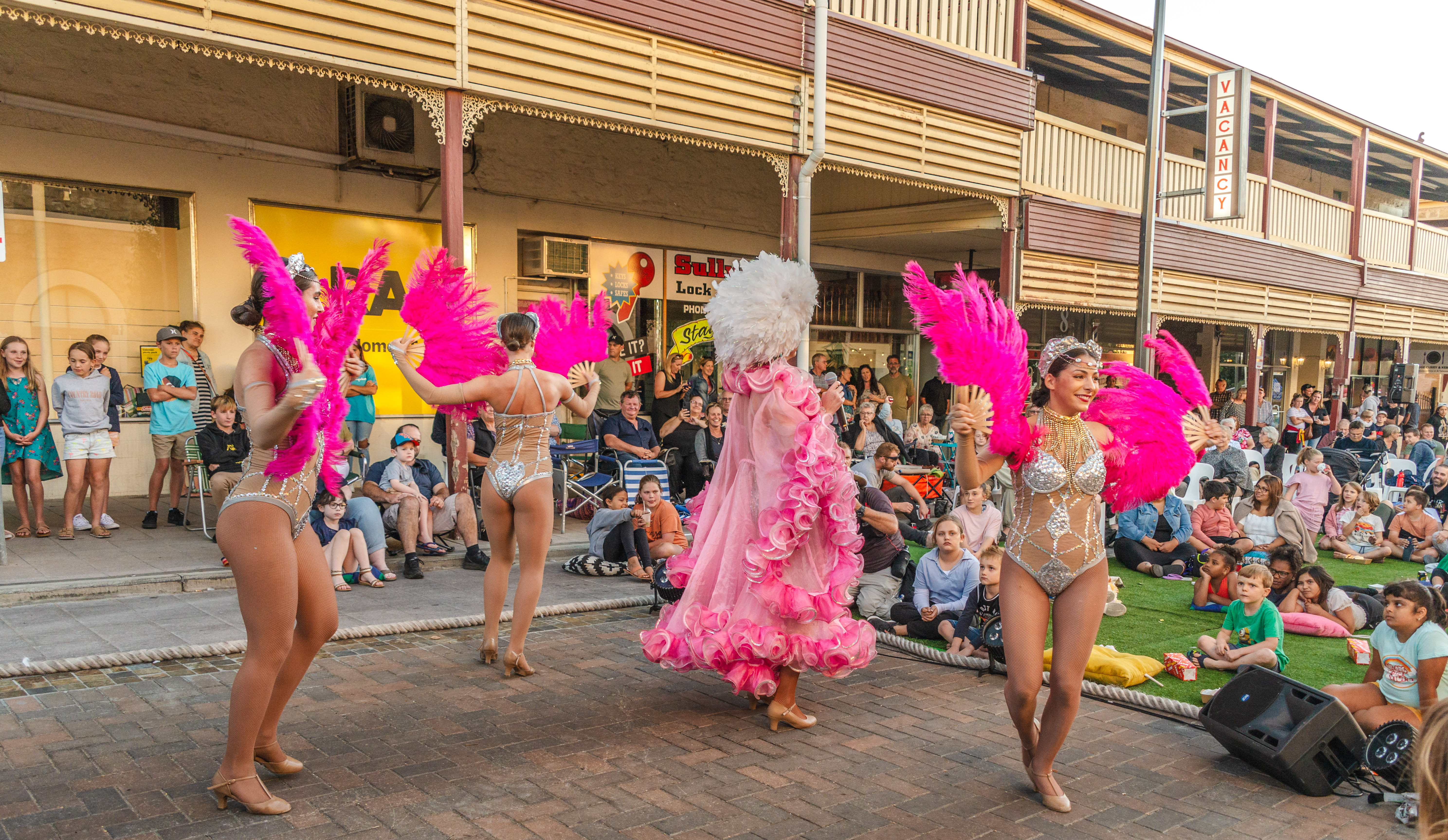 women in colourful costumes holding pink feather fans dance in front of a crowd of people 
