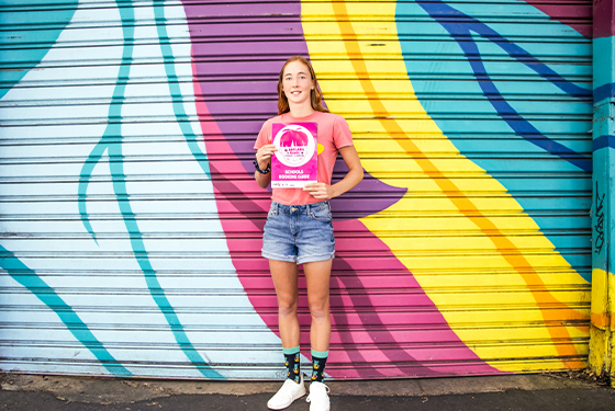 Young girl stands in front of a large painted mural, smiling as she holds a 2022 Adelaide Fringe Schools Guide