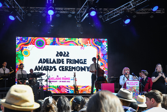 An image of the 2022 Adelaide Fringe Awards Ceremony at The Moa, Gluttony. There is a screen presenting all the Award Winners on the stage. The CEO of Adelaide Fringe, Heather Croall is standing in front of the microphone and giving her speech. One of the ambassadors, Reuben Kaye, musicians and Auslan interpreter, is on the side of the stage.