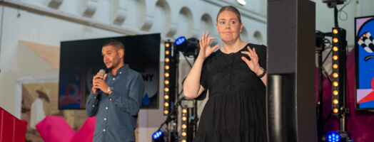 a auslan interpreter at the front of a stage, interpreting the words of a person speaking into a microphone behind them