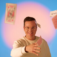 A man throwing playing cards at the camera, illuminated by a large circle peach light in front of a bright blue background