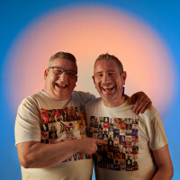 two older men facing the camera in an embrace, illuminated by a peach circle light in front of a bright blue background