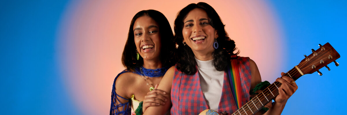 two women in fun clothing, one holding a guitar, smiling at the camera, illuminated by a large peach circle light in front of a bright blue background