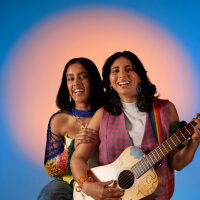 two women in fun clothing, one holding a guitar, smiling at the camera, illuminated by a large peach circle light in front of a bright blue background