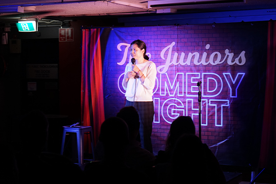 A comedian stands on the stage with purple stage light in the background. 