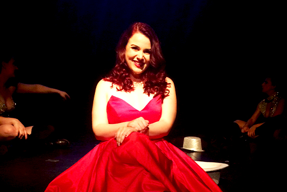 A woman wears a bright red dress sitting in the edge of the stage smiling at the camera. 