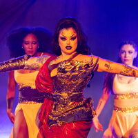 performer in dark makeup with hands outstretched in front of four backup dancers, all standing dramatically in front of a purple backdrop