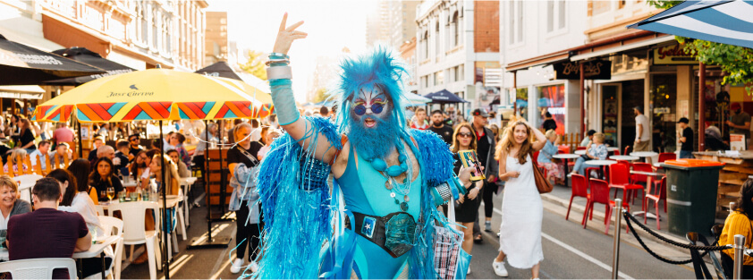 A drag queen standing in the middle of a busy pedestrianised street, dressed head to toe in blue clothes, accessories, hair and makeup
