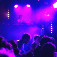 crowd of people dancing to a DJ on a stage in dark purple and pink lighting
