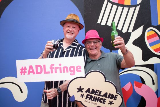 Two smiling men wearing hat infront of a colourful background holding Adelaide Fringe signs