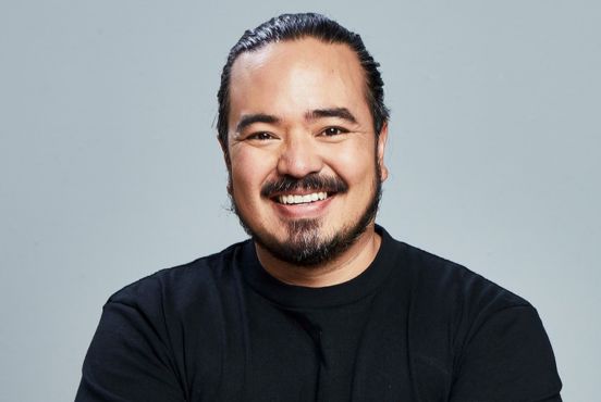 Adam Liaw smiling at the camera, wearing a black tshirt against a grey background