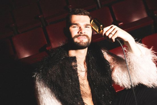 A spotlight illuminates artist Isaac Humphries. He is wearing a black and white feather jacket, holding a golden microphone to his forehead.