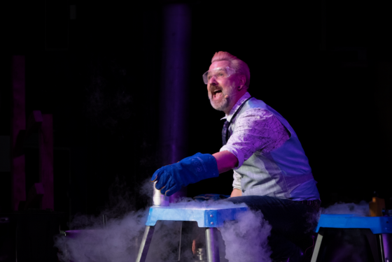 Man wearing protective glasses and gloves holding a tin on a bench with dry ice blowing around the stage