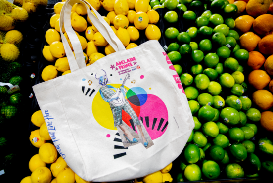A Foodland x Fringe tote bag with a cat playing a guitar on it, situated on top of the lemons, limes and oranges in the fruit and veg section of Foodland
