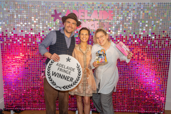 Three people standing in front of a glitter wall. The man on the left is wearing a hat and holding a laurel