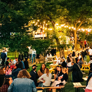 Bustling outdoor evening atmosphere with string lights above, people mingling, dining at picnic tables, and browsing stalls under a canopy of trees.