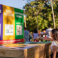 People at an outdoor event putting rubbish into a waste sorting stations labelled 'REUSABLE,' 'MIXED RECYCLING,' and 'COMPOSTABLE ORGANICS' under a canopy of string lights at dusk.