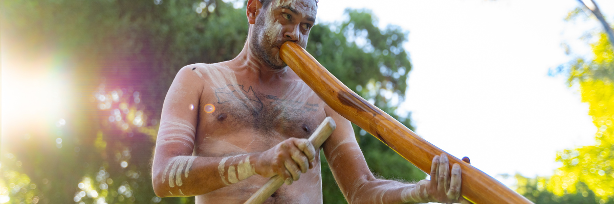 A man with traditional body paint playing the didgeridoo, wearing a red cloth around his waist, with trees softly blurred in the background.