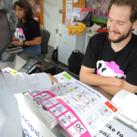 Smiling staff member at the box office with a koala tshirt, assisting a customer with a communication board, while other staff work in the background.
