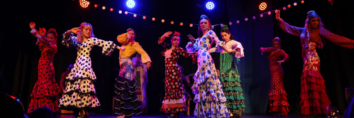 Flamenco dancers in colorful polka-dotted dresses performing on stage with expressive poses under festive lights.