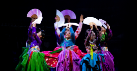 Group of artist on stage with arms outstretched towards the sky. They are all dressed in intricate costumes with vibrant colours.