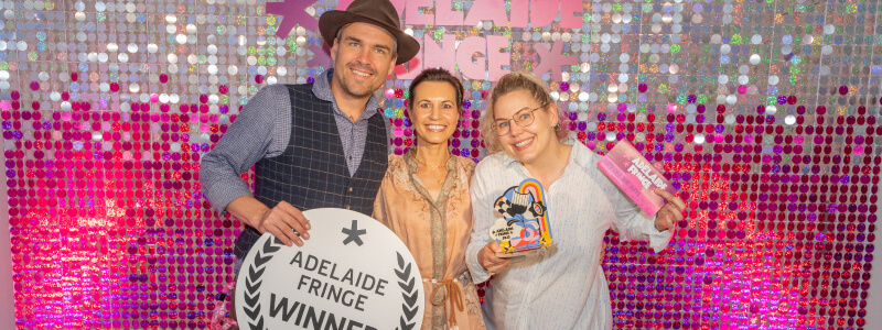three people in an embrace holding awards and signs saying "winner" in front of a silver and pink glittery backdrop