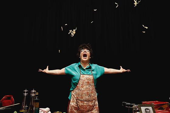 Adelaide Fringe artist, Nella is throwing pasta in the air. Behind her is a black backdrop. 