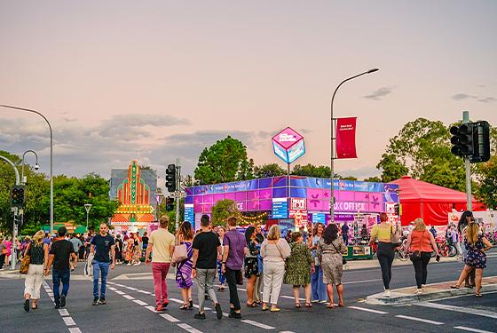 Crowd of people gather and walk across road at twilight in front of purple, pink and blue Adelaide Fringe Corner Box office square structure.  Beside, to the right, a red tent, teetops behind in the distance. 