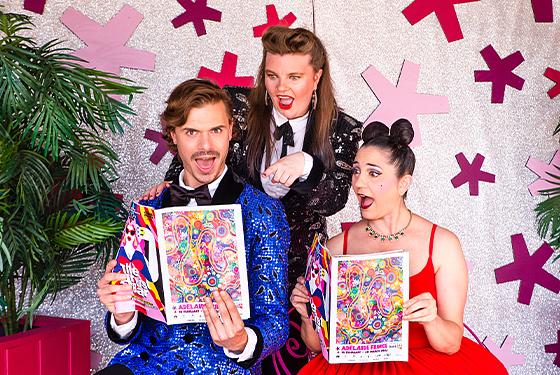 Three performers holding this years Fringe Guide. They are wearing bright sequin clothes and all look exteremly excited. The two on the end are wearing large pink feather headpieces. 