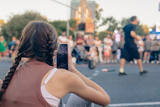 A girl with her phone is photographing a street performer who is juggling. 