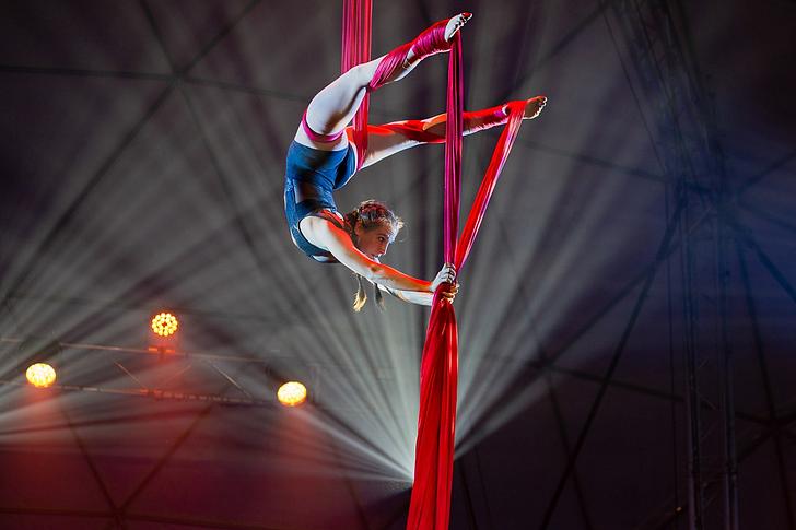 An aerial artist uses red silks to do a backbend stretch above the ground. 