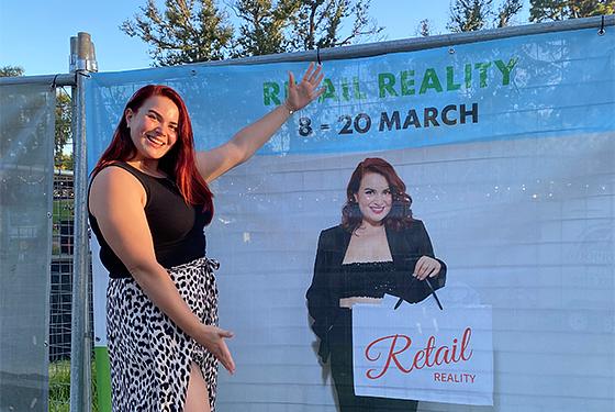 A lady with red long hair stands near a poster with her face on it. She smiles at the camera joyfully. 