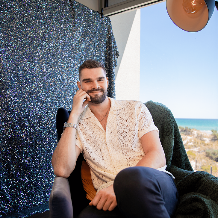 Isaac Humphries sitting on a chair in front of a sequin background and window