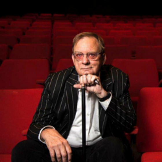 Peter Goers sitting in a theatre with red seats, resting his chin on a walking stick