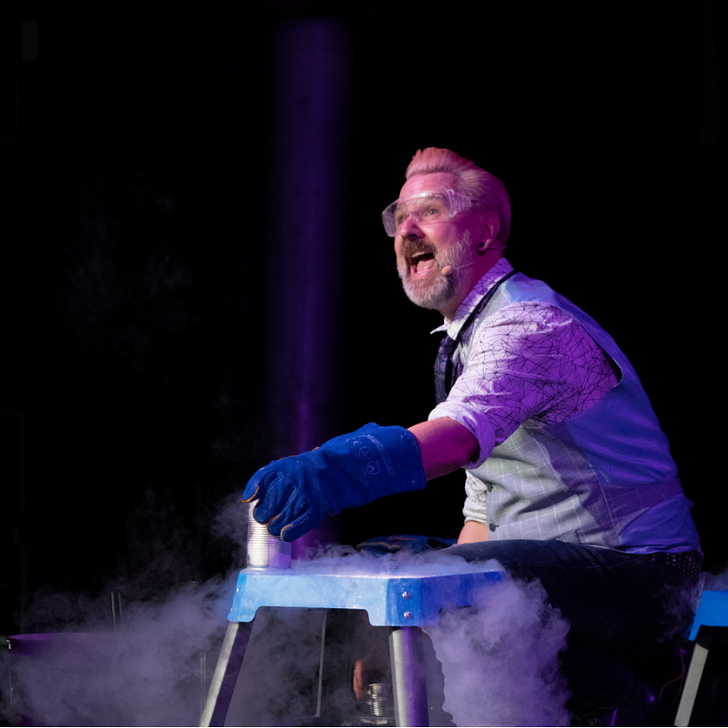 Man wearing protective glasses and gloves holding a tin on a bench with dry ice blowing around the stage