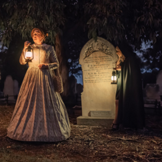 Two women in 1800s outfits holding lanterns in a cemetery 