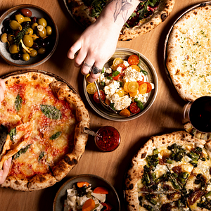 Close up of a table covered in food including pizzas with someone reaching towards the food