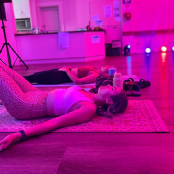 Two ladies are lying down on the floor and relaxing with their eyes closed. The room is dimly lit, with pink lights.